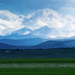 Snow-capped Rockies above green fields in Severance, Colorado, under a cloudy sky.