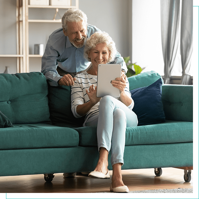 Senior Couple Enjoying Technology: Visually Impaired Accessible Description of Elderly Pair Using Tablet on Sofa Senior couple smiling while using a tablet together on a teal sofa in a well-lit living room, depicting technology use across generations.